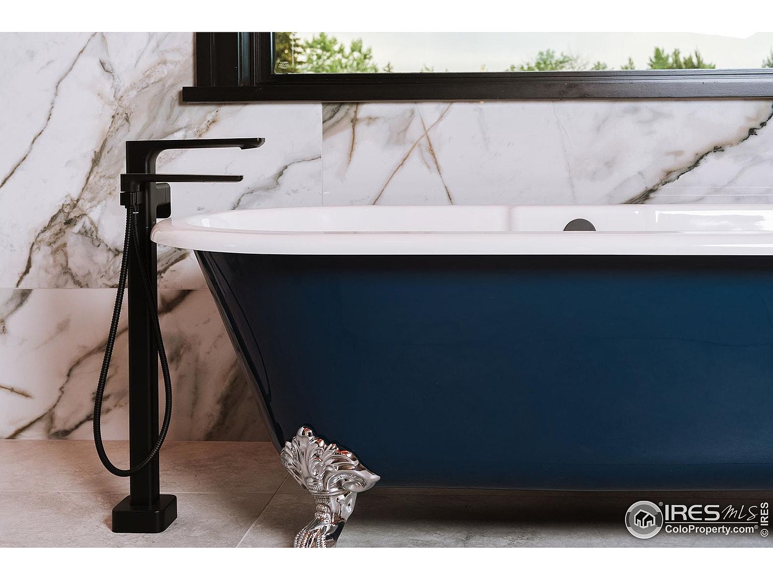 This image showcases a luxurious primary bathroom featuring a freestanding, deep blue bathtub with ornate silver claw feet. A modern, matte black faucet stands beside the tub, complementing the marble-tiled walls. The overall impression is one of elegance and relaxation, perfect for a high-end real estate listing.