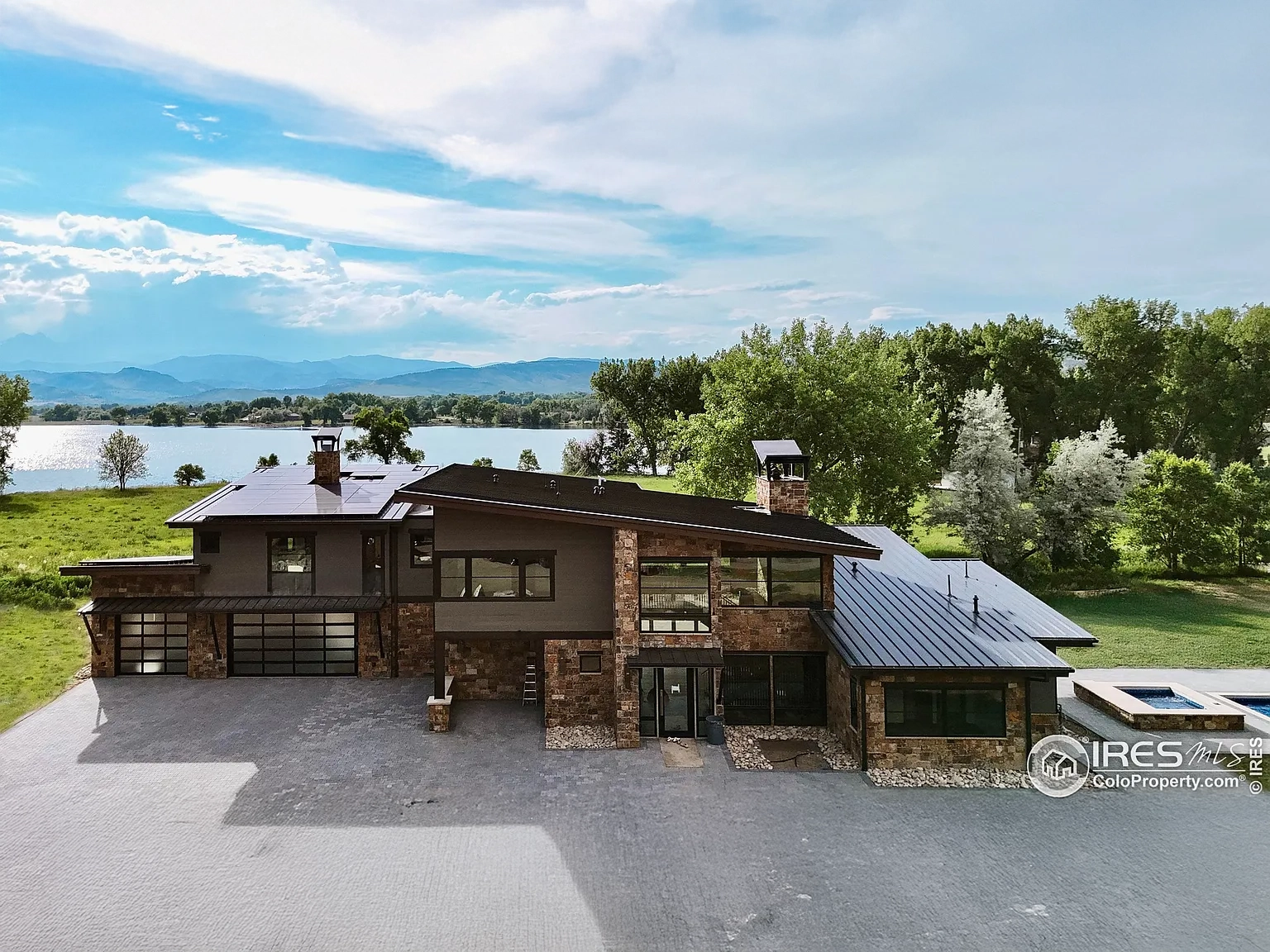 This is an aerial view of a contemporary home situated near a lake or body of water. The house features a combination of stone and modern siding, a dark metal roof, and a multi-car garage. A pool or spa is visible in the yard, contributing to a luxurious and private feel.