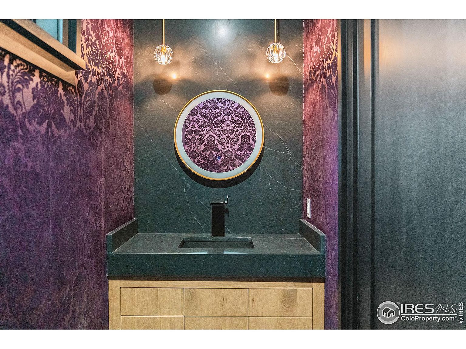 This is a stylish guest bathroom featuring a round mirror with a gold frame, mounted above a dark countertop and light wood vanity. The walls are adorned with a combination of dark marble-like tile and patterned purple wallpaper, complemented by modern pendant lighting. The overall design creates a sophisticated and luxurious feel.