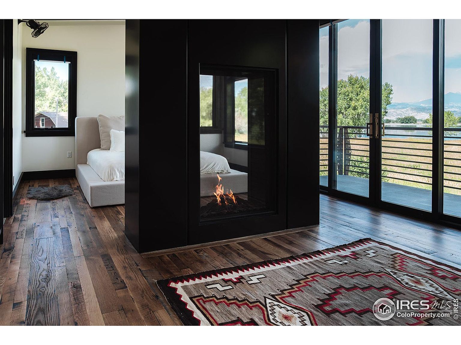 This is an interior shot of a primary bedroom featuring a modern fireplace that divides the sleeping area from a balcony. The room has hardwood floors and a neutral color palette, with a bed visible on both sides of the fireplace. Large sliding glass doors lead to the balcony, offering a view of the landscape.