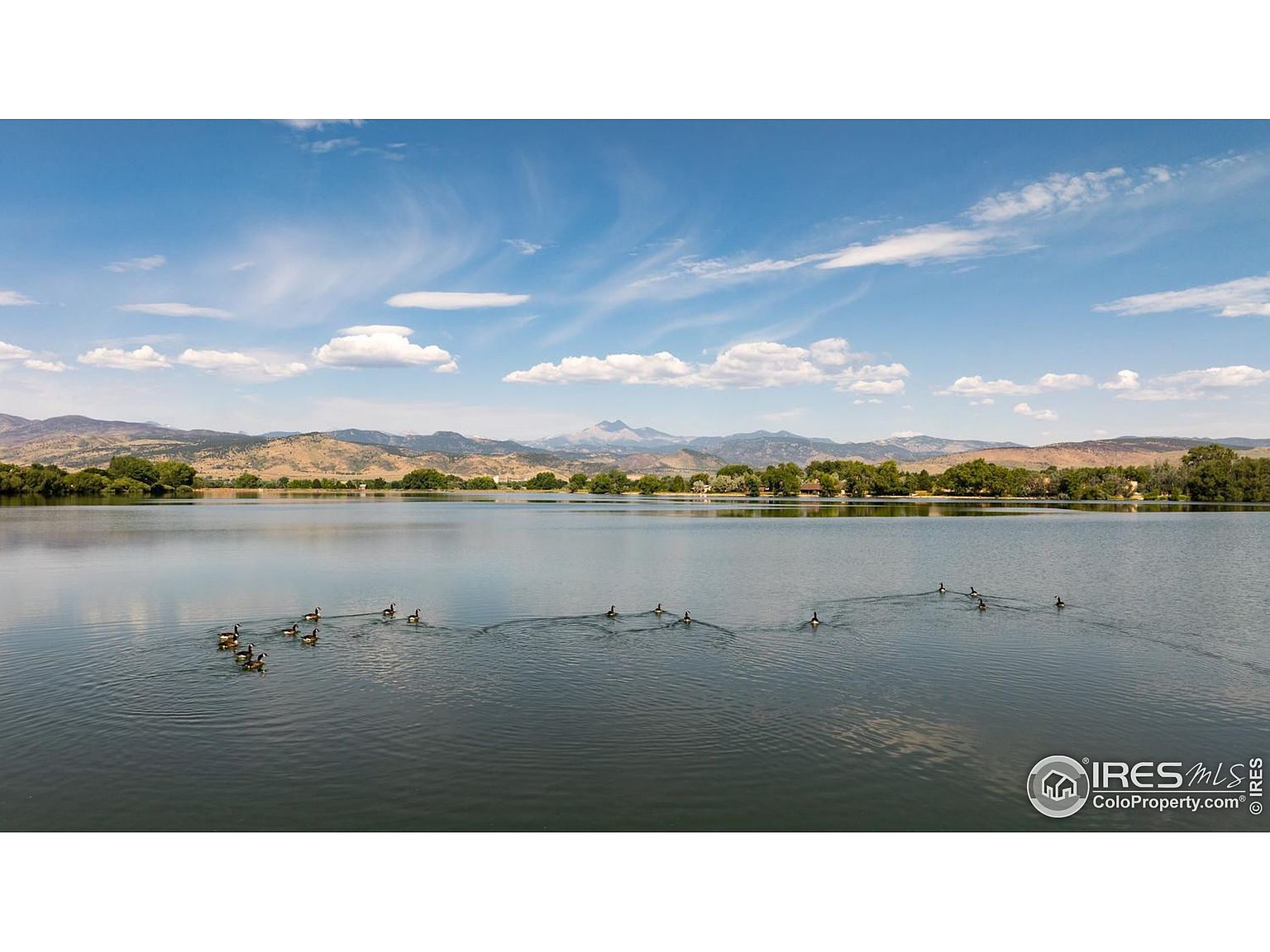 This image showcases a serene lake scene, likely part of a community amenity. The calm water reflects the blue sky and scattered clouds, with a backdrop of rolling hills and mountains. A group of ducks swims across the lake, adding to the peaceful atmosphere.