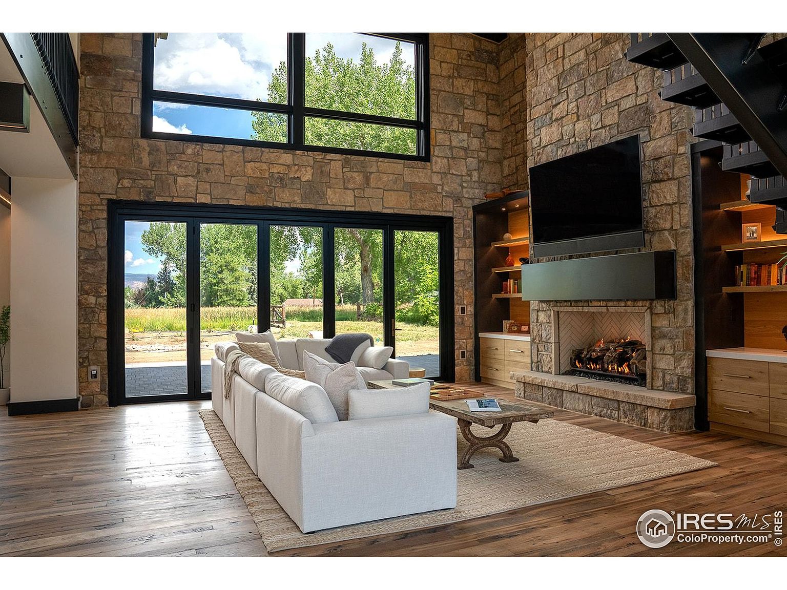 This is an interior shot of a living room featuring a stone fireplace with a mounted television, built-in shelving units, and a large sectional sofa. The room has a high ceiling with a large window and sliding glass doors that offer a view of the outdoors. The hardwood flooring and neutral color palette create a warm and inviting atmosphere.