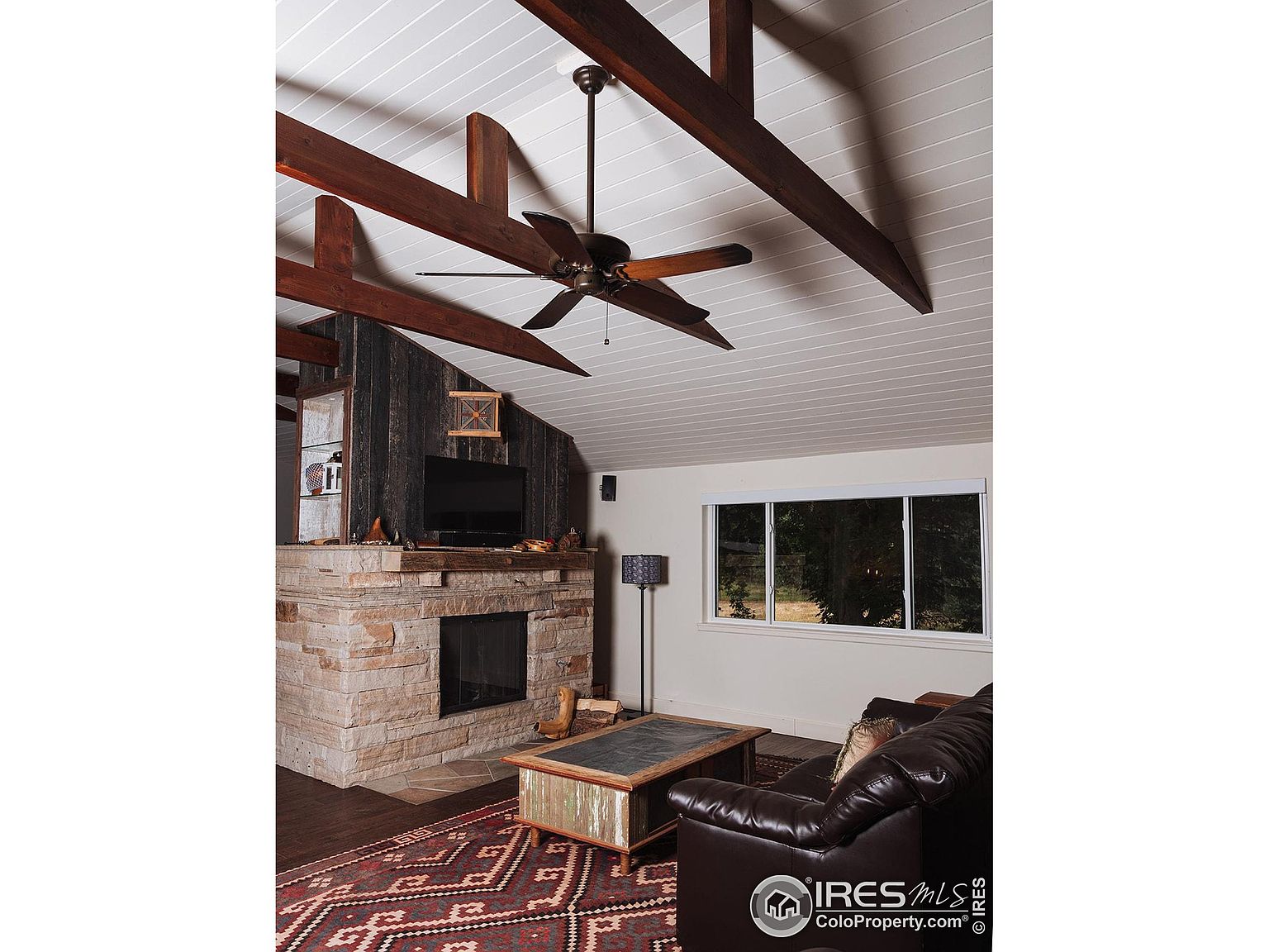 This is an interior shot of a living room featuring a stone fireplace with a dark wood mantle and a television mounted above. The room has exposed wooden beams on the ceiling and a large window providing natural light. A leather sofa and a patterned rug add to the cozy and rustic atmosphere of the space.
