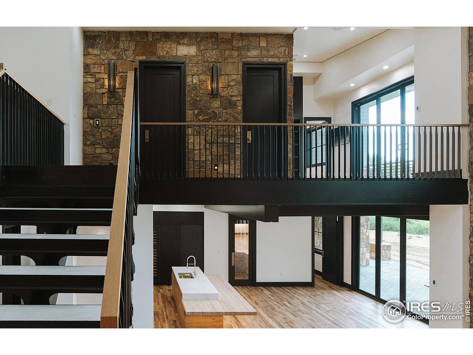 This interior shot showcases a modern home's hallway and staircase. The design features a stone accent wall on the upper level with two dark doors, complemented by a black railing and wooden stairs. The open floor plan allows for a view into the kitchen area, highlighting the home's spaciousness and contemporary style.