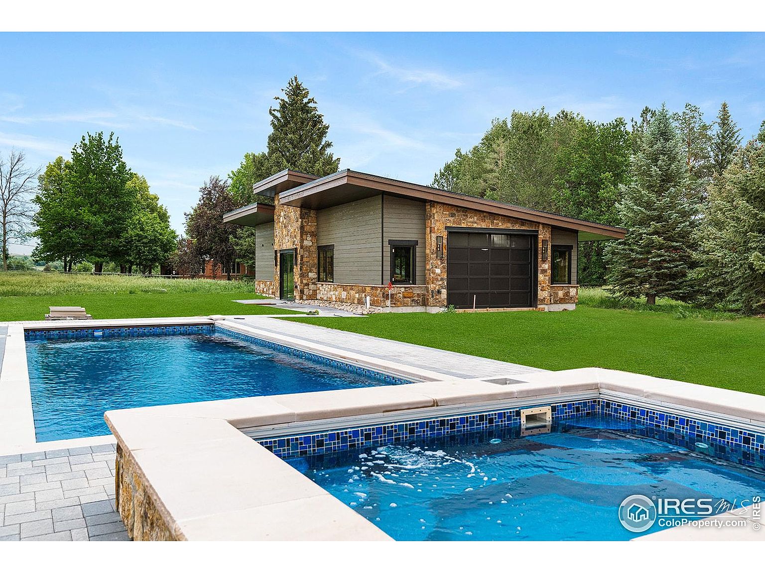 This image showcases a luxurious outdoor pool and spa area. The pool features clear blue water and is complemented by a spa with blue mosaic tiles. The surrounding landscape includes a well-maintained lawn and a modern house with a garage, creating an inviting and upscale atmosphere.