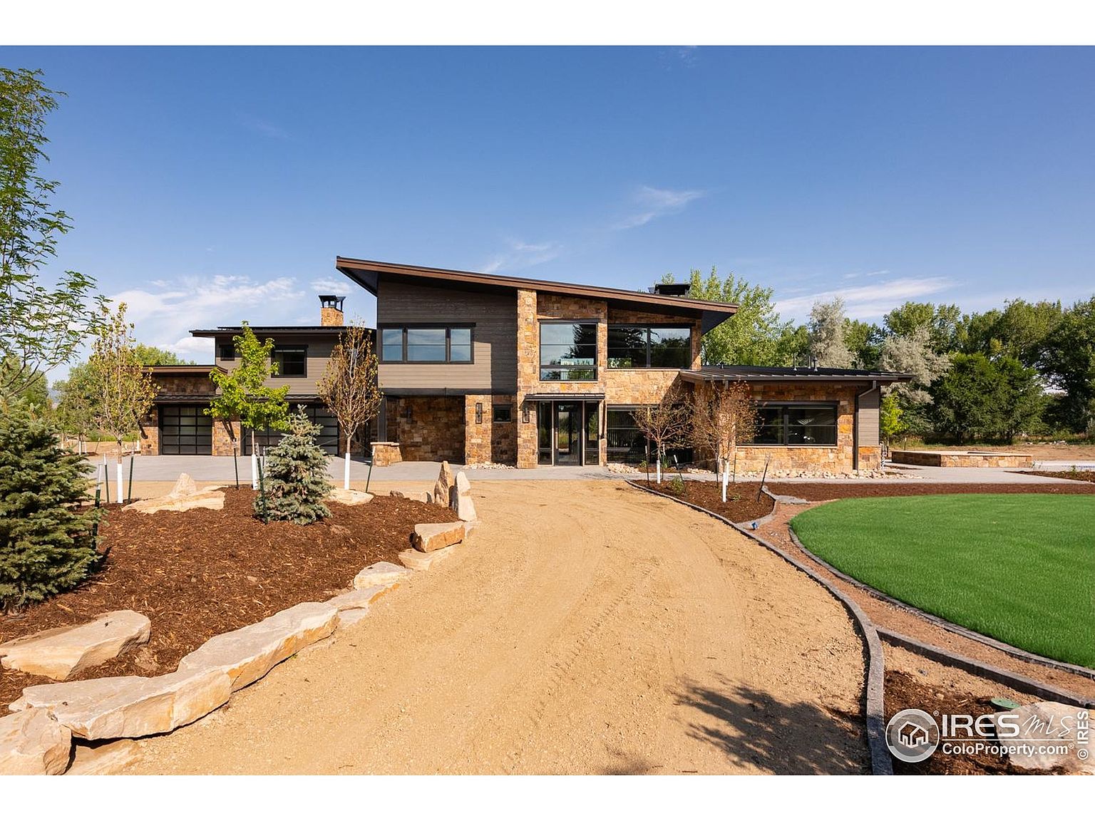 This is a front exterior view of a modern, two-story home with a unique roofline. The facade features a combination of stone and wood siding, large windows, and a well-manicured front yard with a circular driveway. The landscaping includes mature trees, shrubs, and decorative rock features, enhancing the property's curb appeal.