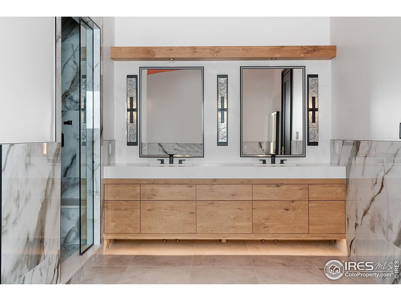 This is a well-lit primary bathroom featuring a double vanity with a white countertop and light wood cabinetry. Two rectangular mirrors are mounted above the sinks, flanked by decorative sconces. A glass-enclosed shower is visible to the left, and the walls are adorned with marble-like tiles, creating a luxurious and modern aesthetic.