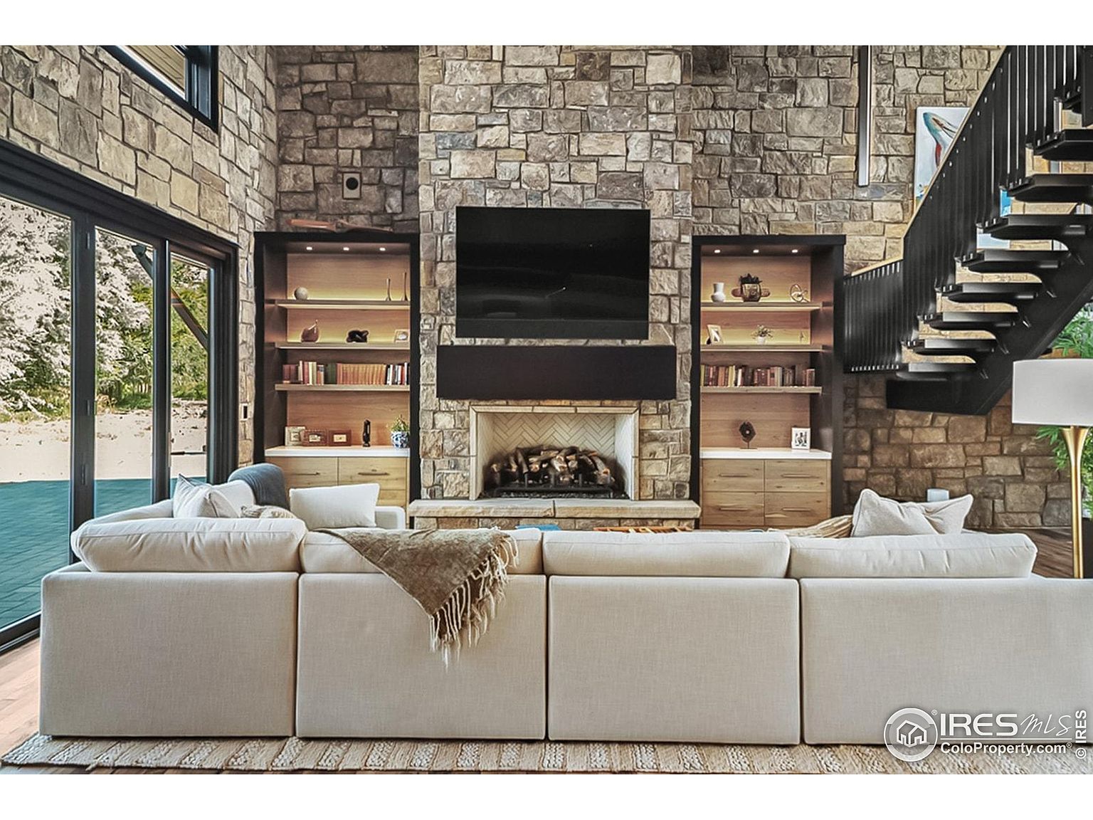 This is an interior shot of a living room featuring a large sectional sofa in a neutral color, positioned in front of a stone fireplace with a mounted television. Built-in bookshelves flank the fireplace, and a modern staircase is visible on the right. The room has a contemporary design with a focus on natural materials and textures, creating a warm and inviting atmosphere.