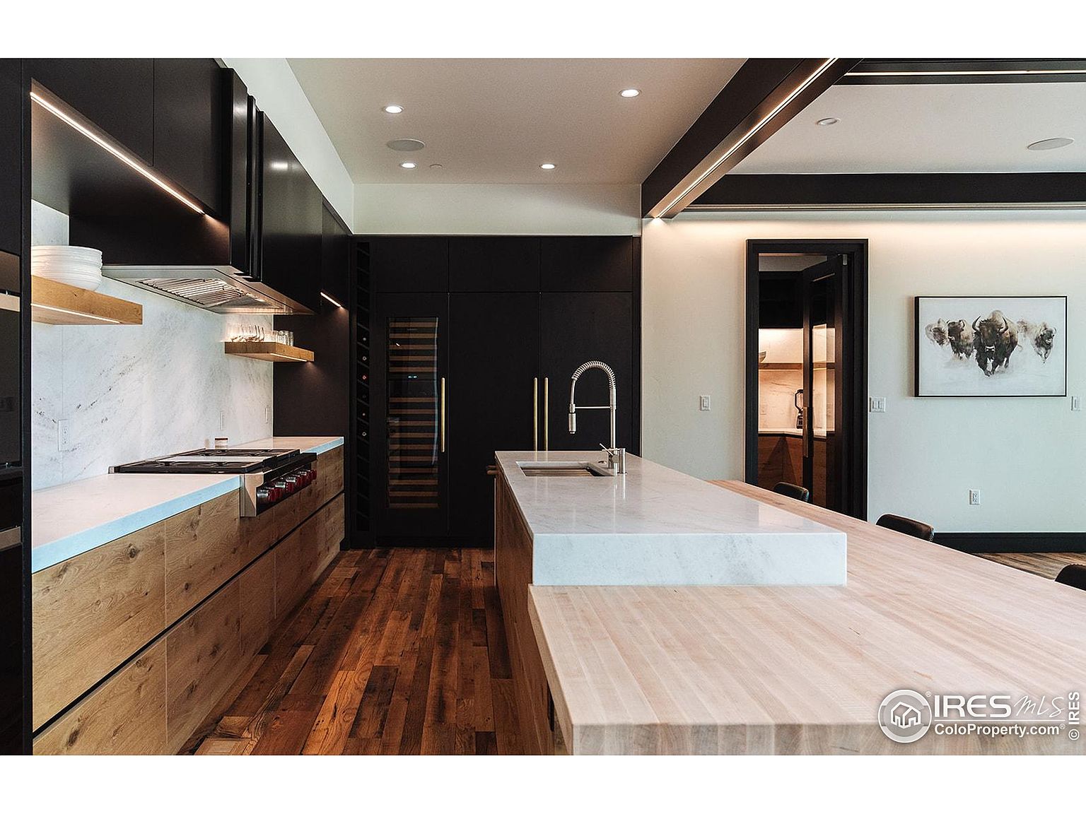 This is a modern kitchen with a blend of dark and light wood cabinetry, complemented by white countertops and marble backsplash. The kitchen features a large island with a sink, a built-in wine cooler, and stainless steel appliances. The hardwood flooring adds warmth to the space, while the black accents provide a contemporary contrast.