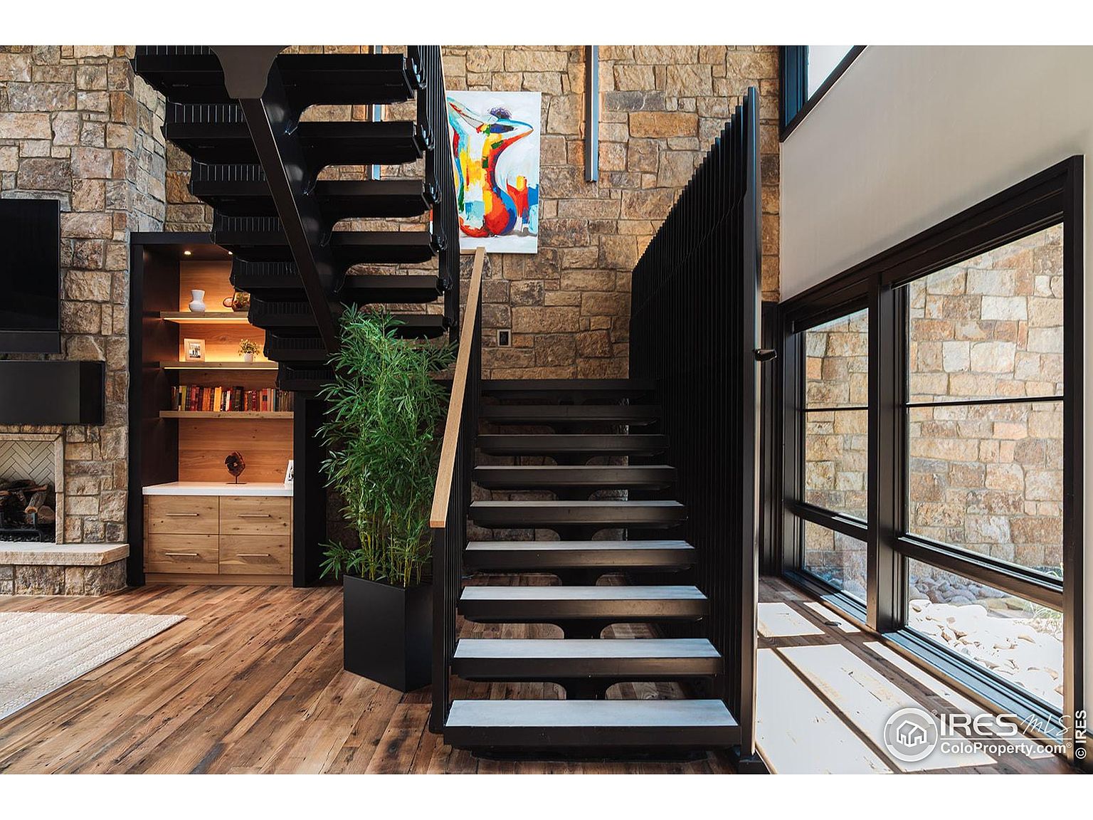 This interior shot showcases a modern staircase with open treads and a black metal railing, complemented by a natural wood handrail. The staircase is flanked by a stone wall and a large window, providing ample natural light. A vibrant abstract painting adds a pop of color to the space, while a potted plant brings a touch of nature indoors.