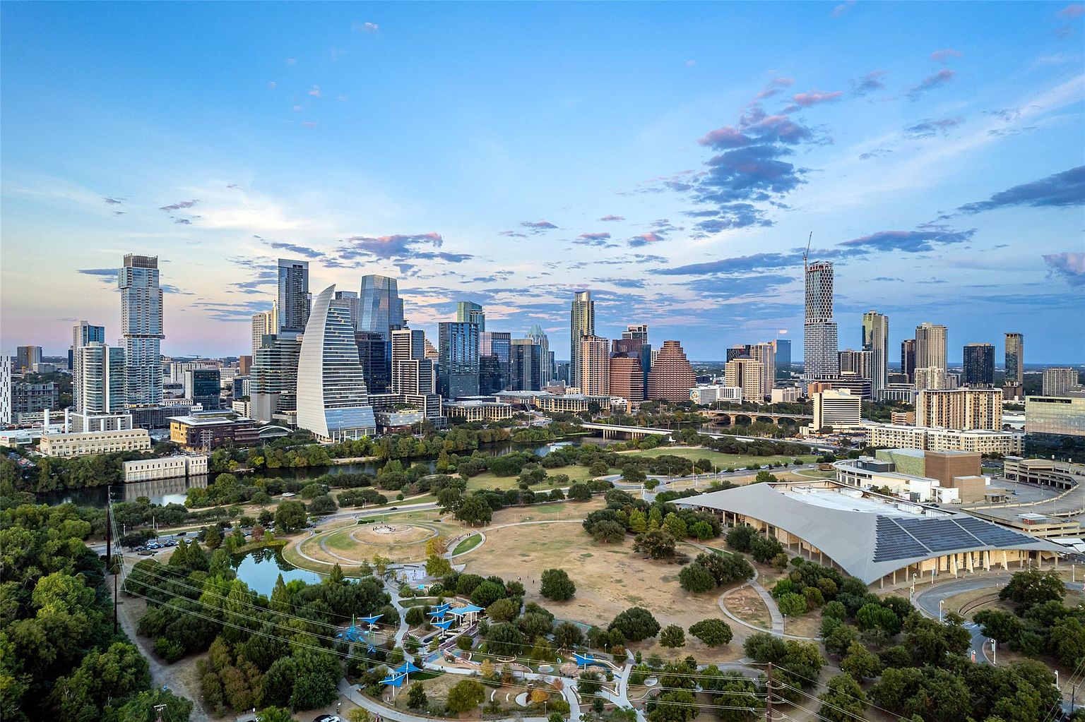 This aerial view showcases the Austin, Texas skyline, featuring modern skyscrapers and architectural landmarks against a backdrop of a partly cloudy sky. Below, a lush green park with a pond and walking paths adds a natural element to the urban landscape. The image captures the vibrant energy and scenic beauty of the city, highlighting its blend of urban development and green spaces.