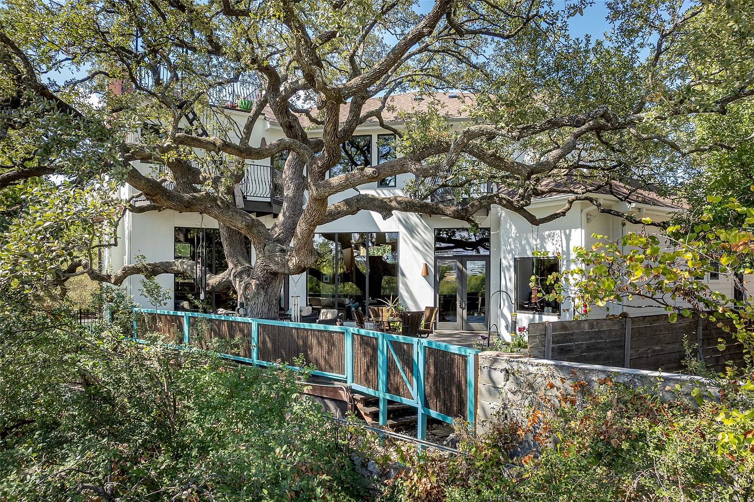This is a rear view of a two-story white house, partially obscured by a large, mature tree with sprawling branches. The house features large windows and a balcony, suggesting an open and airy interior. A wooden deck with a blue railing extends from the house, providing an outdoor living space.
