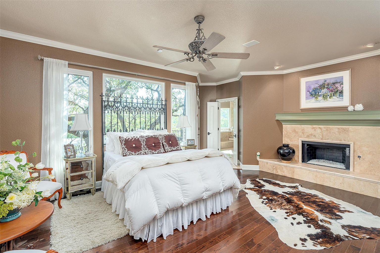 This is a primary bedroom featuring a large bed with white bedding and decorative pillows, a decorative headboard, and a cowhide rug on hardwood floors. The room has a fireplace with a decorative mantel and a painting above it, as well as windows with sheer curtains. The style is traditional and luxurious, creating a cozy and inviting atmosphere.