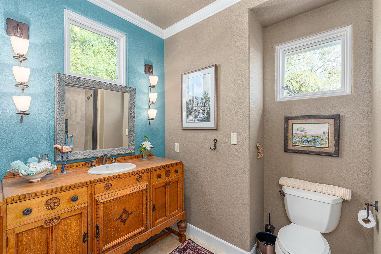 This is a charming guest bathroom featuring a unique, antique-style wooden vanity with an integrated sink. The walls are painted in a two-tone scheme, with a teal accent wall behind the vanity and a neutral beige on the remaining walls. The room is decorated with framed artwork and vintage-inspired light fixtures, creating a cozy and inviting atmosphere.
