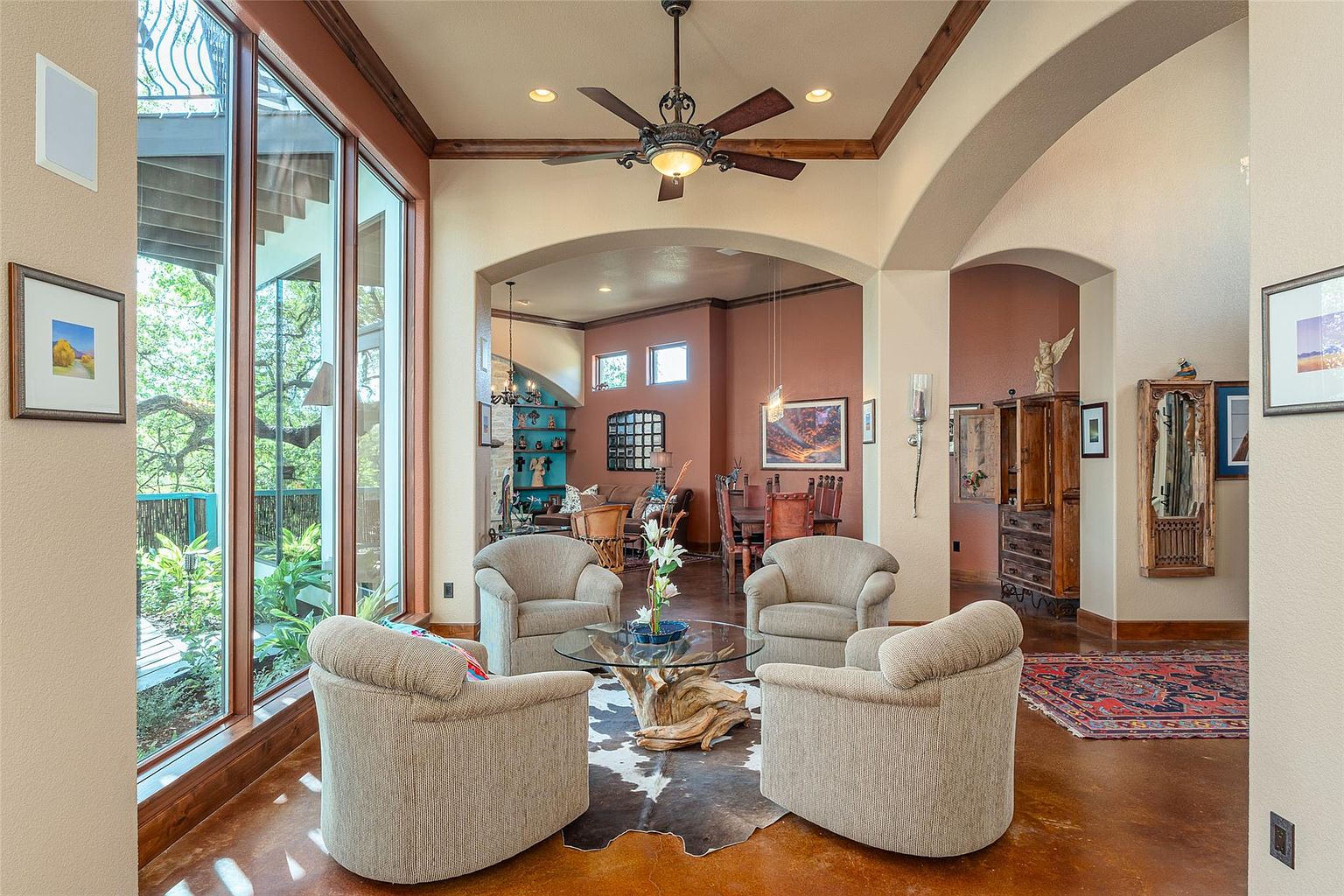 This is an interior shot of a living room featuring a seating area with four armchairs arranged around a unique glass-topped coffee table with a driftwood base, set on a cowhide rug. The room is open to adjacent spaces through arched doorways, and a large window provides natural light and a view of the outdoors. The style is rustic-chic with warm tones and wooden accents.