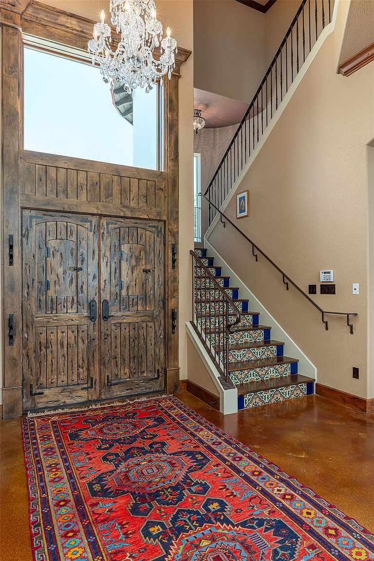 This interior shot showcases an elegant entryway featuring a grand wooden double door and a staircase with decorative tile risers. A vibrant, patterned rug adds warmth to the polished floor, while a chandelier hangs above, illuminating the space. The overall impression is one of rustic luxury and inviting sophistication.