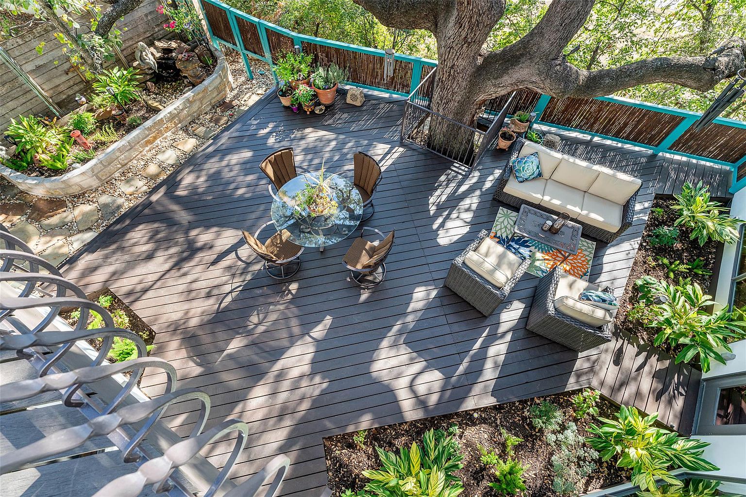 This is an aerial view of a stylish outdoor deck area. The deck features a dining set with a glass table and four chairs, a comfortable seating area with a sofa and two armchairs, and integrated planters with lush greenery. The space is designed around a large tree, creating a serene and inviting atmosphere.