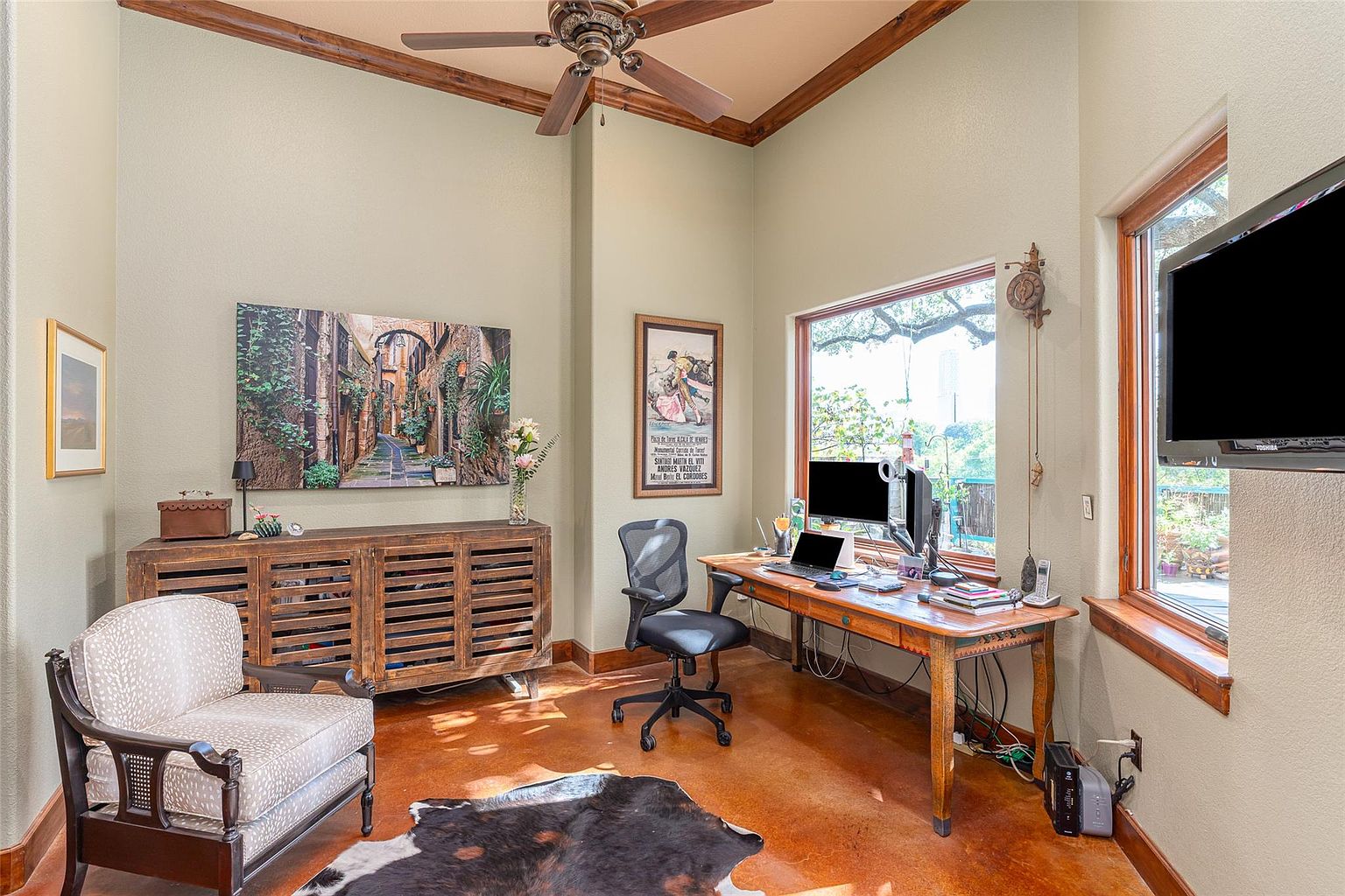 This is an interior shot of a home office featuring a wooden desk with a computer setup, a comfortable office chair, and a decorative cabinet with artwork above. The room has a warm and inviting feel, with natural light streaming in through a window, complemented by a ceiling fan and a unique cowhide rug on the floor. A television is mounted on the wall near the window.