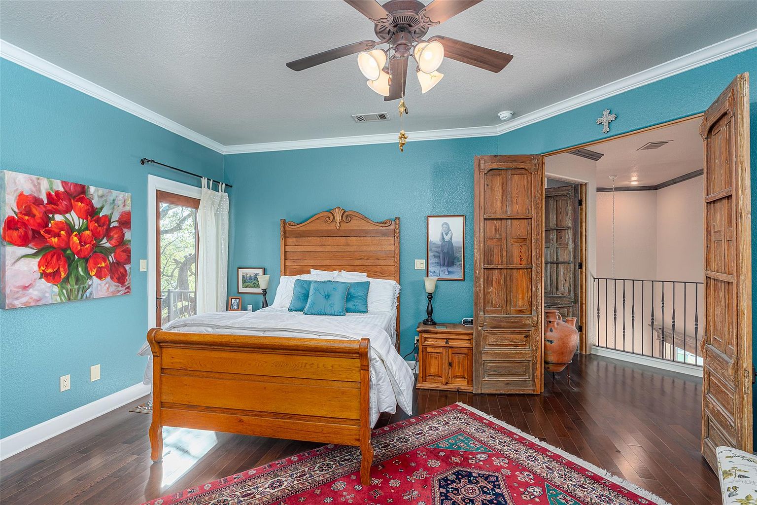 This is a primary bedroom featuring a wooden sleigh bed with blue accent pillows, a red and blue patterned rug, and hardwood floors. The walls are painted a calming blue, complemented by white crown molding. The room opens to a second-floor landing through rustic wooden doors, adding a unique architectural element.