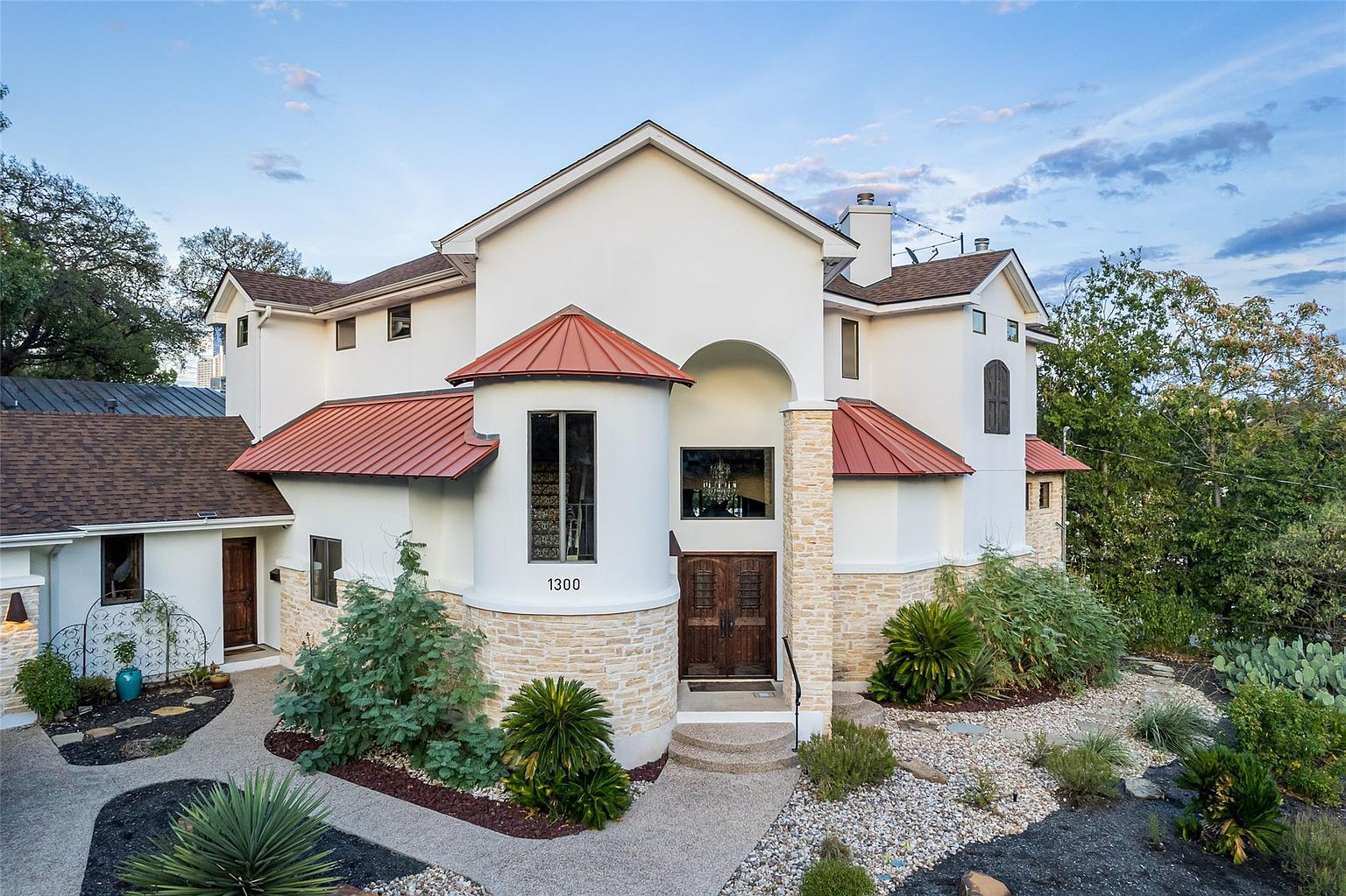 This is a front exterior view of a two-story house with a unique architectural design. The house features a combination of white stucco and stone accents, with a red metal roof adding a pop of color. The landscaping is well-maintained with a mix of plants, gravel, and stone pathways leading to the front entrance, creating an inviting curb appeal.