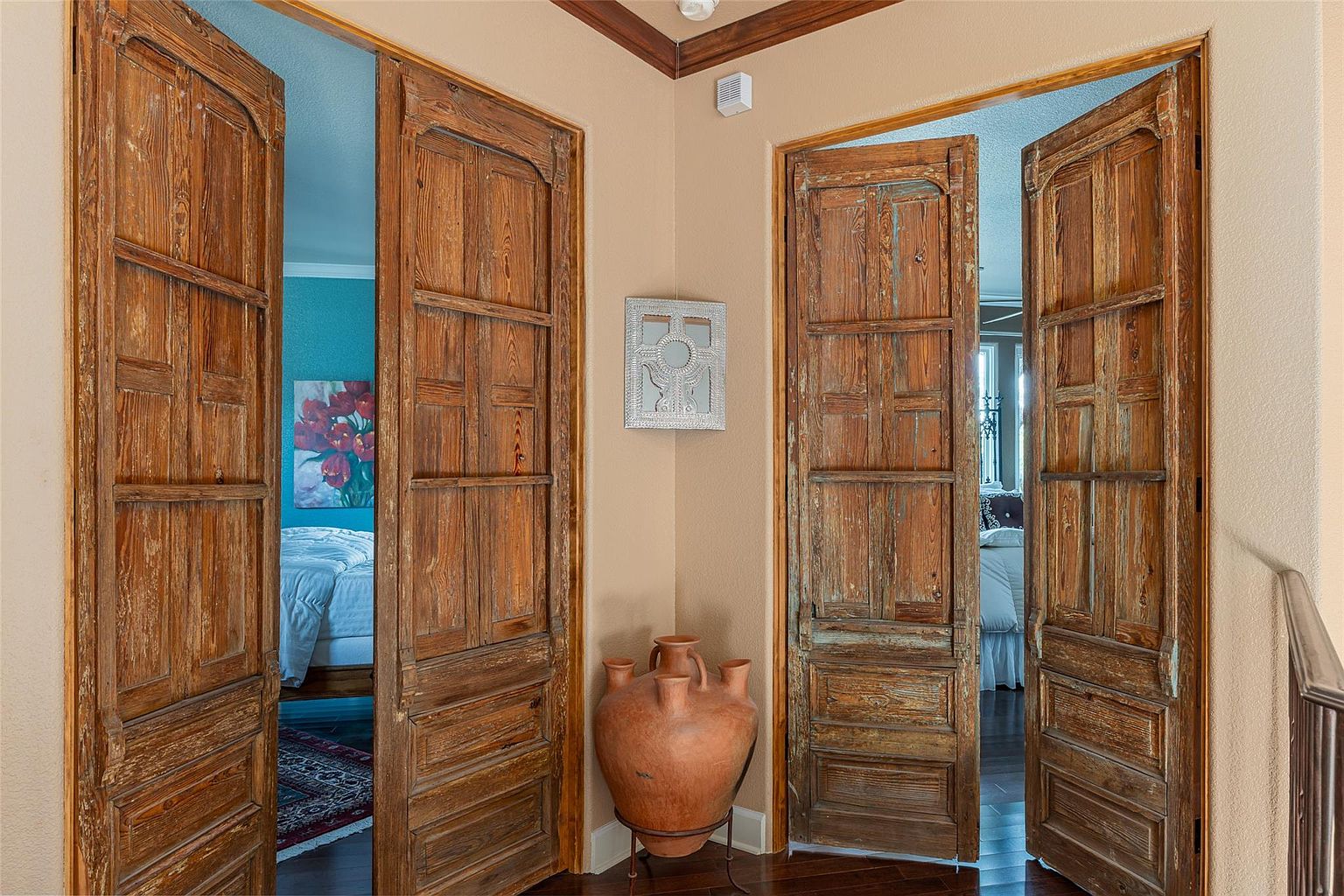 The image showcases a hallway featuring two sets of rustic, wooden double doors leading into bedrooms. A large, decorative clay pot sits in the corner, adding to the home's character. The warm color palette and unique architectural details create an inviting and stylish transitional space.