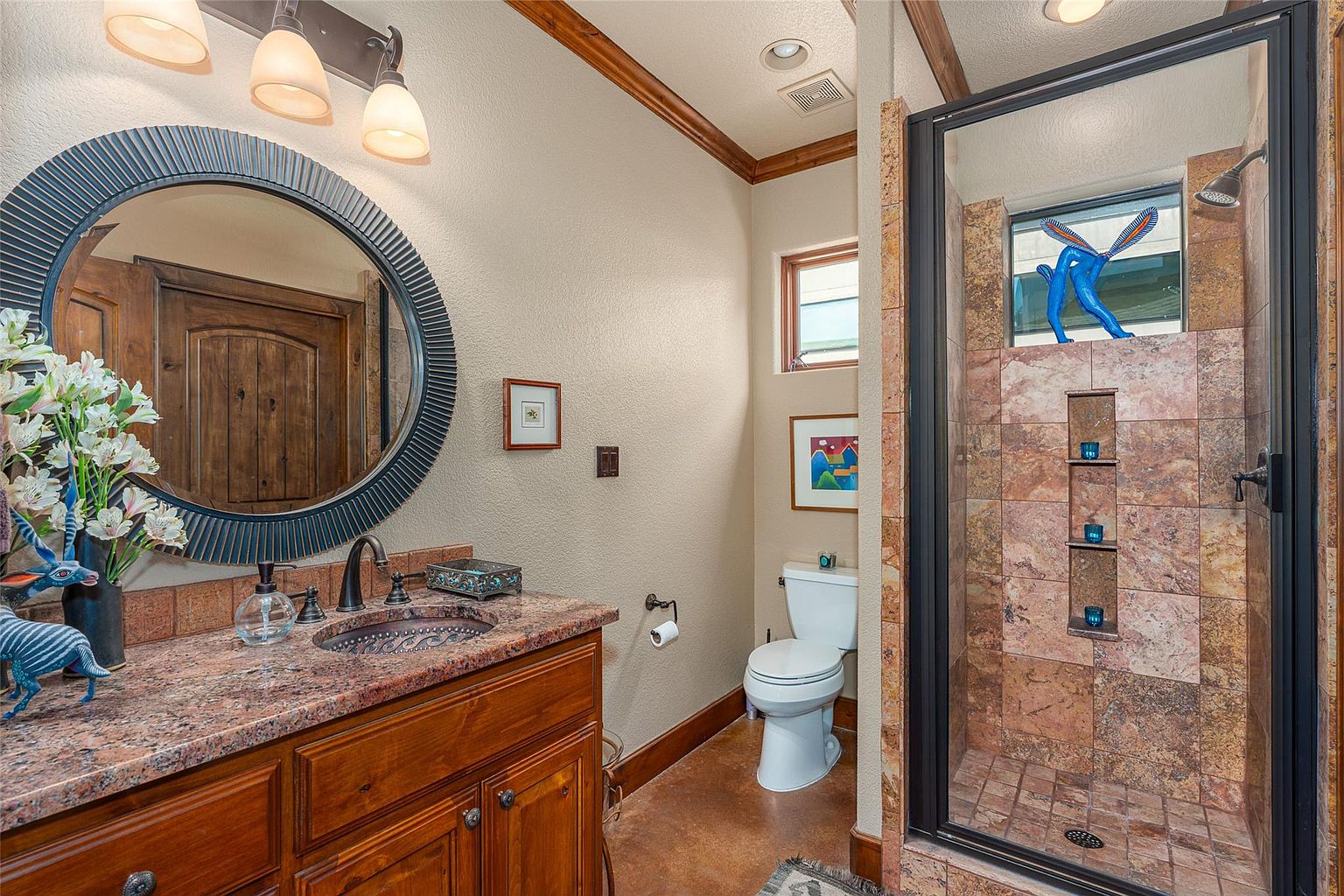 This is a well-appointed guest bathroom featuring a granite-topped vanity with a copper sink and a round mirror. The shower is enclosed with glass and features decorative tilework and built-in shelving. The room has a warm, inviting feel with earth-toned walls and wooden trim.