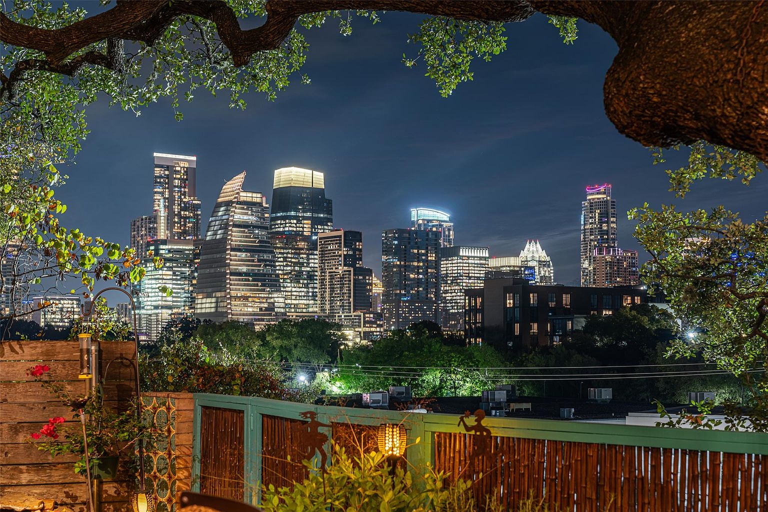 This image showcases a charming patio or balcony with a stunning city skyline view at night. The foreground features a wooden fence adorned with decorative metal cutouts and a bamboo screen, adding a touch of privacy and character. Lush greenery and a tree branch frame the scene, creating a cozy and inviting outdoor space perfect for enjoying the illuminated cityscape.