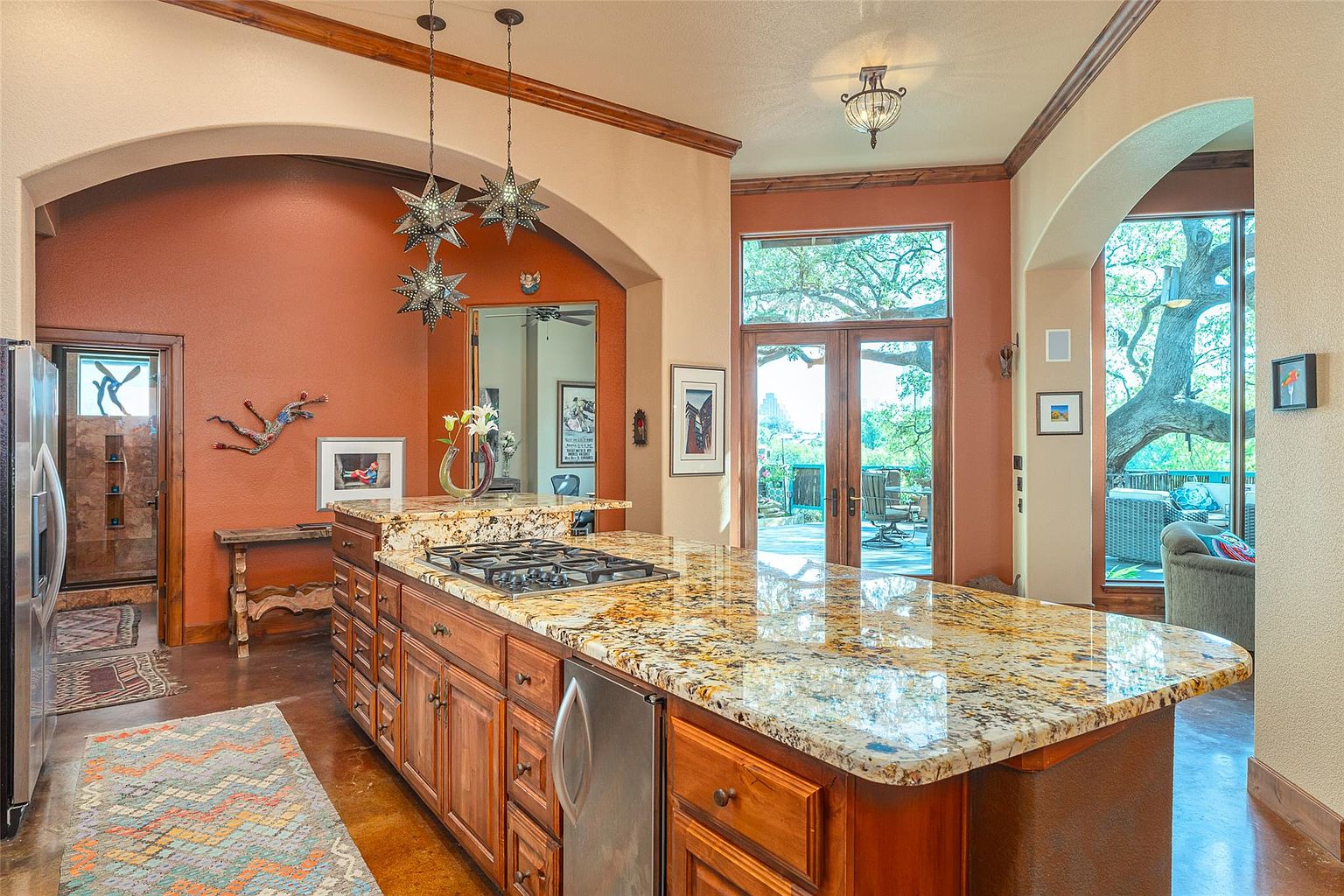 This is a warm and inviting kitchen featuring a large island with a granite countertop and a built-in cooktop. The cabinetry is a rich wood tone, and the walls are painted in earthy colors. The kitchen has an open feel with archways leading to other areas of the home and a large window/door combination providing natural light and views of the outdoors.