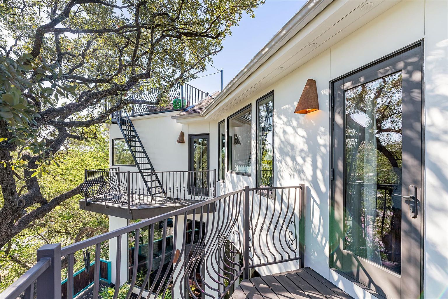 This image showcases a multi-level patio/deck/balcony area of a modern home, featuring wrought iron railings and a unique architectural design. The exterior is painted white, and the decks are made of wood. A striking tree is integrated into the design, adding a natural element to the structure.