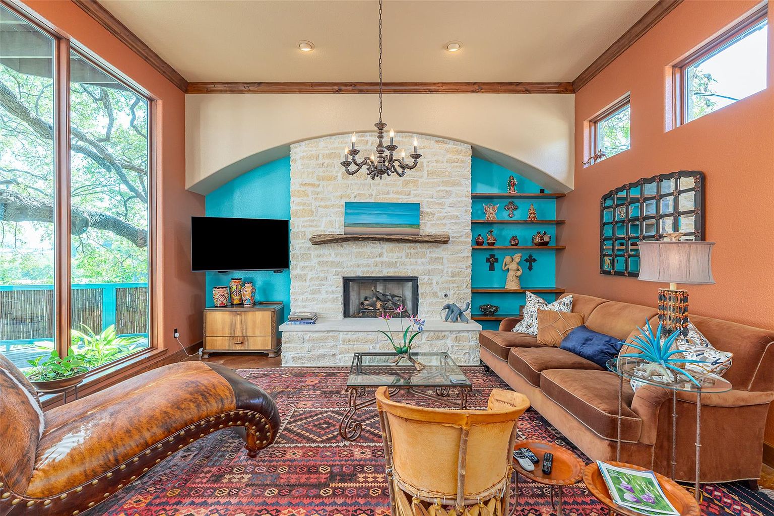 This living room features a stone fireplace with a wooden mantel and built-in shelves, creating a focal point. The room is furnished with a brown sofa, a leather chaise lounge, and a glass-topped coffee table, all set upon a patterned rug. Large windows offer natural light and views of the outdoors, enhancing the room's inviting atmosphere.