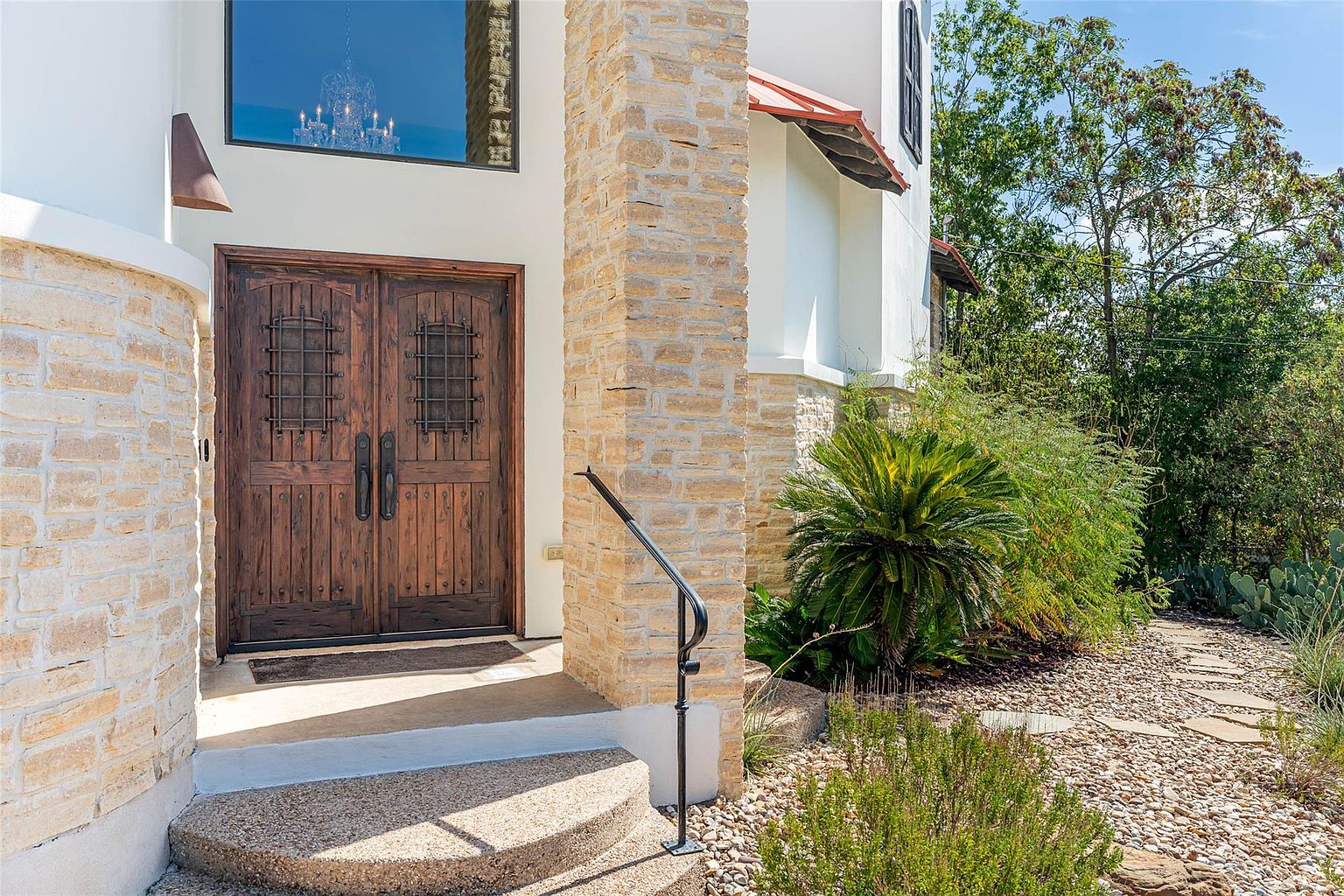This image showcases the entryway of a home, featuring a set of ornate wooden double doors with iron detailing. The entrance is framed by stone pillars and a small set of steps leading up to the door. Landscaping elements, including a small palm tree and gravel pathway, add to the curb appeal, creating a welcoming and sophisticated first impression.
