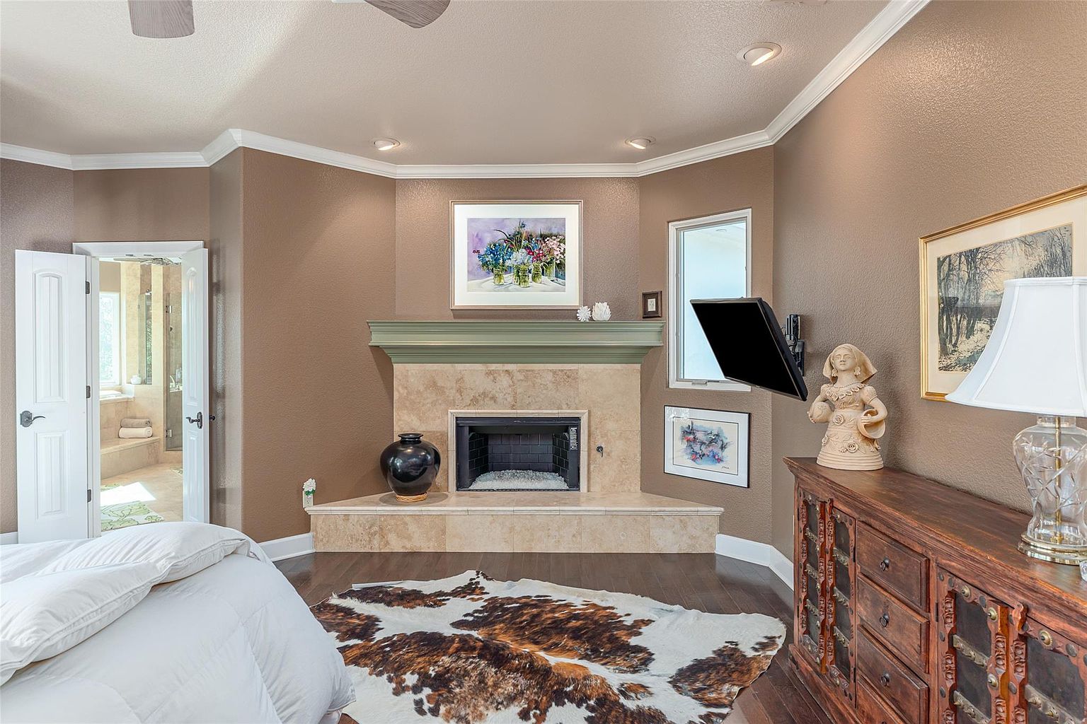 This is a primary bedroom featuring a fireplace with a stone surround and a decorative mantel. The room has a warm color palette with brown walls and white trim. A large dresser with a statue and lamp sits against one wall, and a cowhide rug adds texture to the hardwood floor. A doorway leads to an en-suite bathroom.