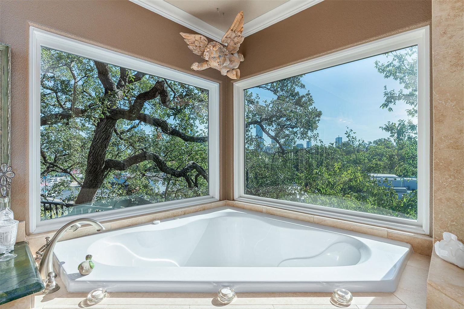 This is a luxurious primary bathroom featuring a large, triangular soaking tub positioned in front of a corner window offering a scenic view of lush trees and a distant cityscape. The bathroom is decorated with neutral tones and features a decorative angel statue above the window, creating a serene and spa-like atmosphere. The perspective is from a medium shot, showcasing the tub and the view.