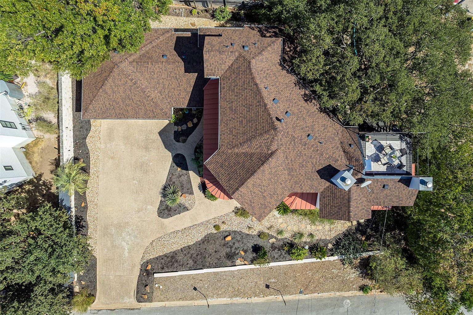 This aerial shot showcases a well-maintained residential property with a brown shingle roof, a spacious driveway, and meticulously landscaped grounds. The house features a unique architectural design with multiple rooflines and a rooftop patio area, surrounded by mature trees that provide shade and privacy. The overall impression is one of a luxurious and private home.