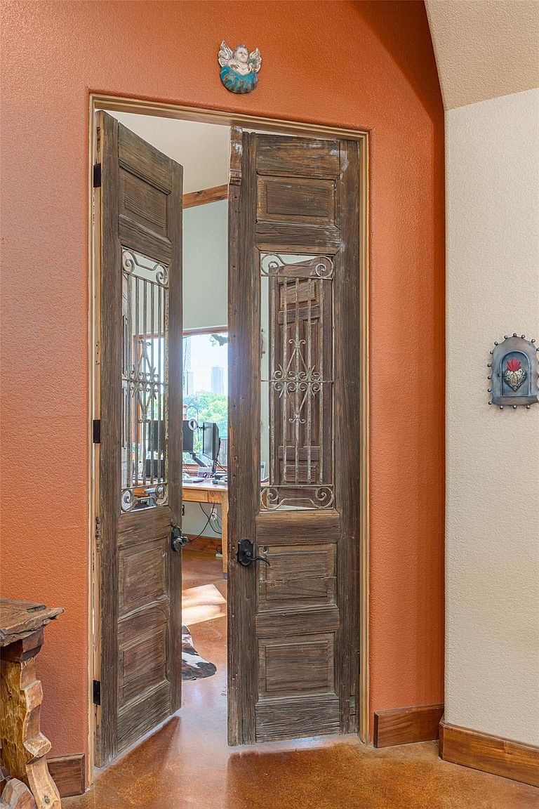 The image showcases an interior hallway featuring rustic wooden double doors with decorative ironwork, partially open to reveal an office space. The walls are textured with a warm, earthy tone, complemented by wooden baseboards and decorative wall art. The flooring appears to be polished concrete, adding to the home's unique character and charm.