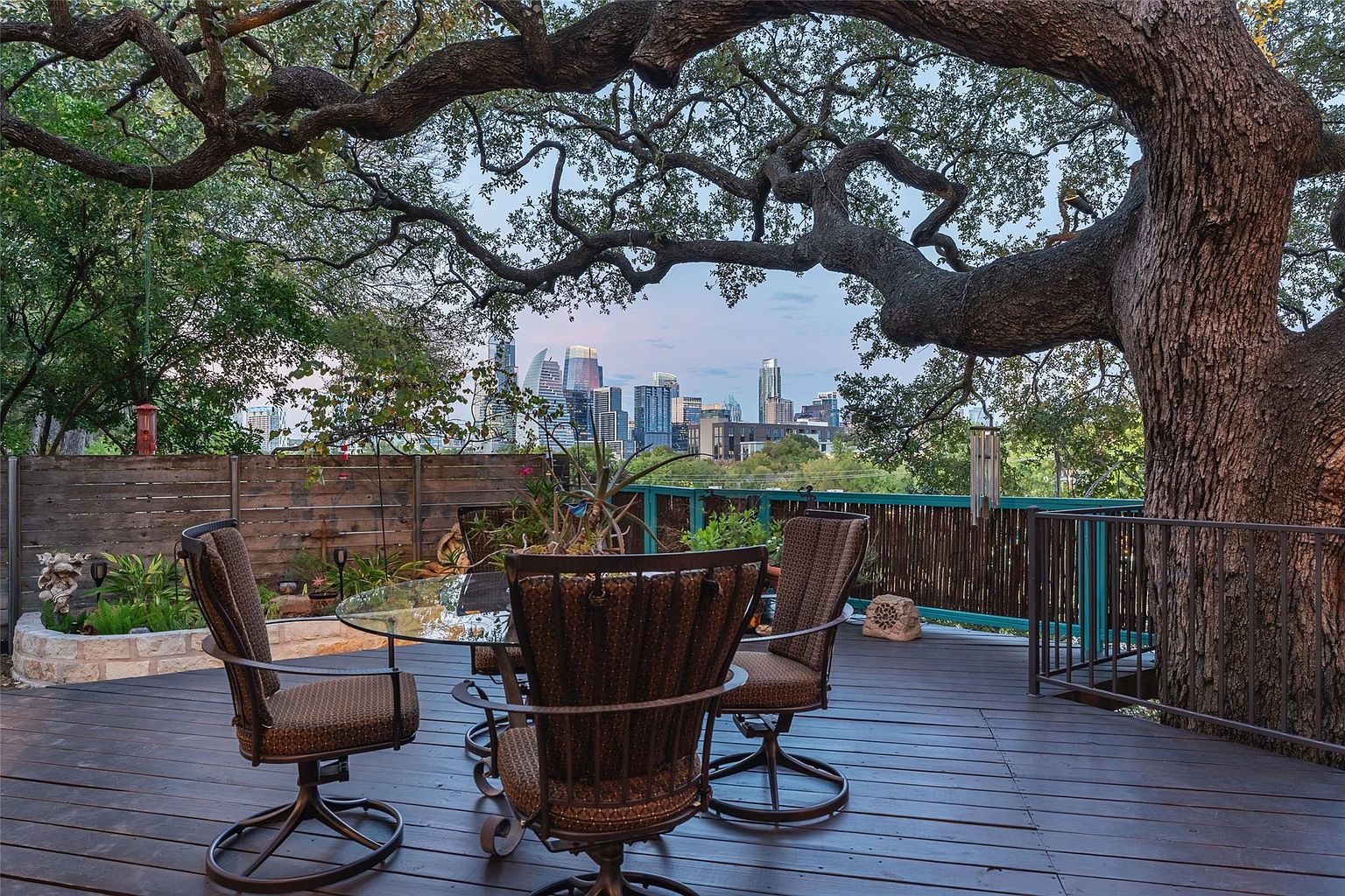 This image showcases a charming outdoor patio or deck area, featuring a round glass-top table surrounded by four wicker chairs. The deck is constructed of dark wood and is partially shaded by a large, mature tree, offering a view of the city skyline in the distance. The setting evokes a sense of relaxation and outdoor living.