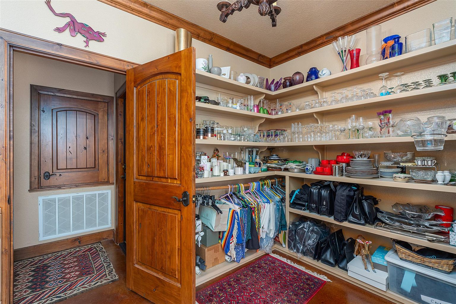 This image showcases a well-organized pantry with wooden shelving stocked with glassware, dishes, and various household items. A wooden door is partially open, revealing a glimpse into an adjacent room. The pantry features a warm color palette and a rustic charm, making it both functional and aesthetically pleasing.