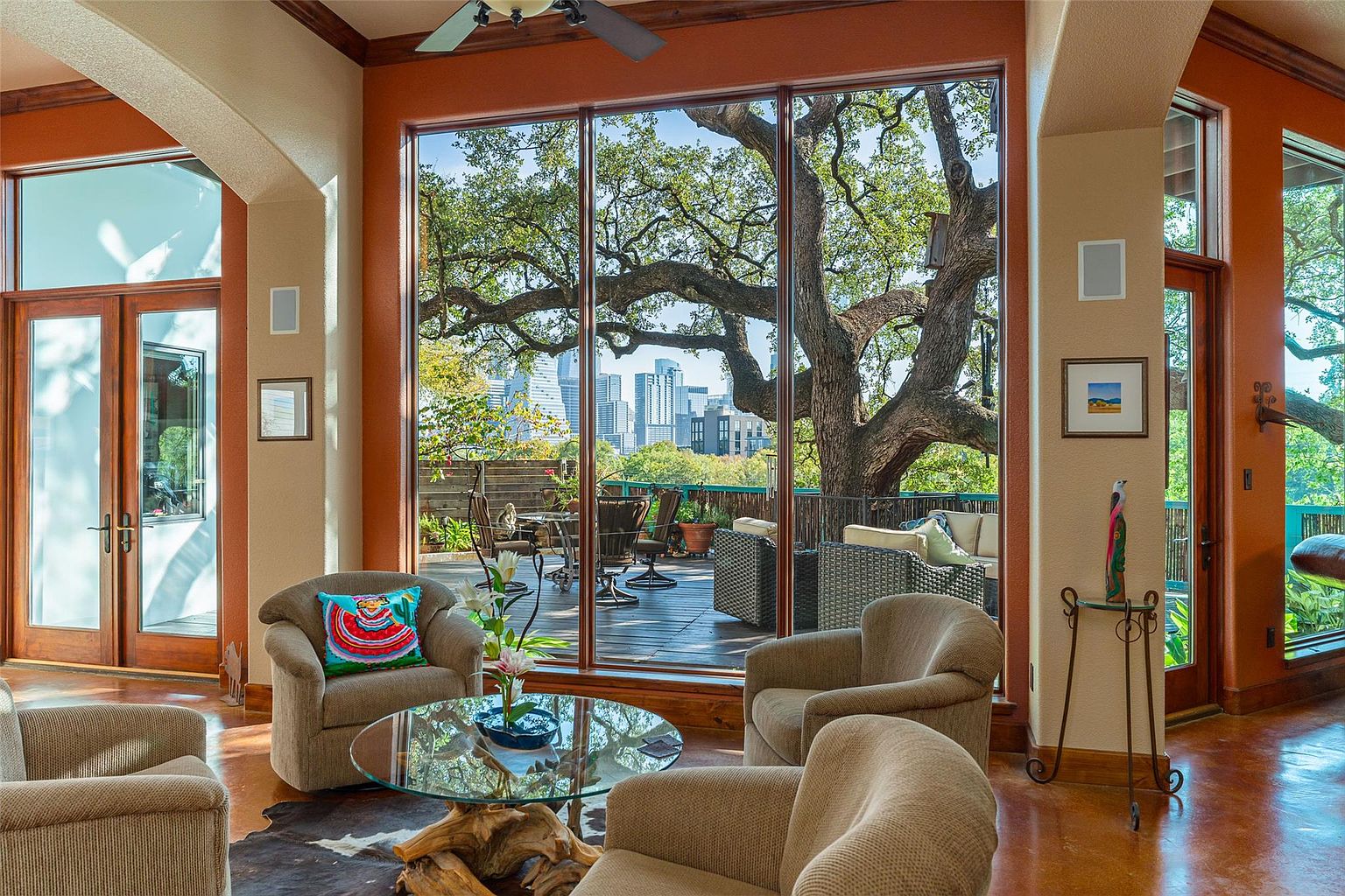This is an interior shot of a living room featuring comfortable armchairs arranged around a glass-topped coffee table with a unique wooden base. Large windows offer a stunning view of a tree and the city skyline, creating a seamless connection between indoor and outdoor spaces. The room is decorated with warm tones and natural materials, giving it a cozy and inviting atmosphere.