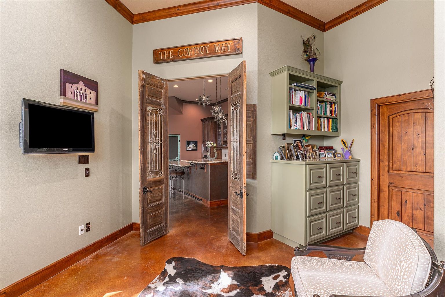 This interior shot showcases a living room with a rustic-chic aesthetic. The room features unique wooden doors leading to another space, a built-in cabinet with shelving, and a cowhide rug on a polished floor. A television is mounted on the wall, and a sign reading 'The Cowboy Way' adds to the room's character.