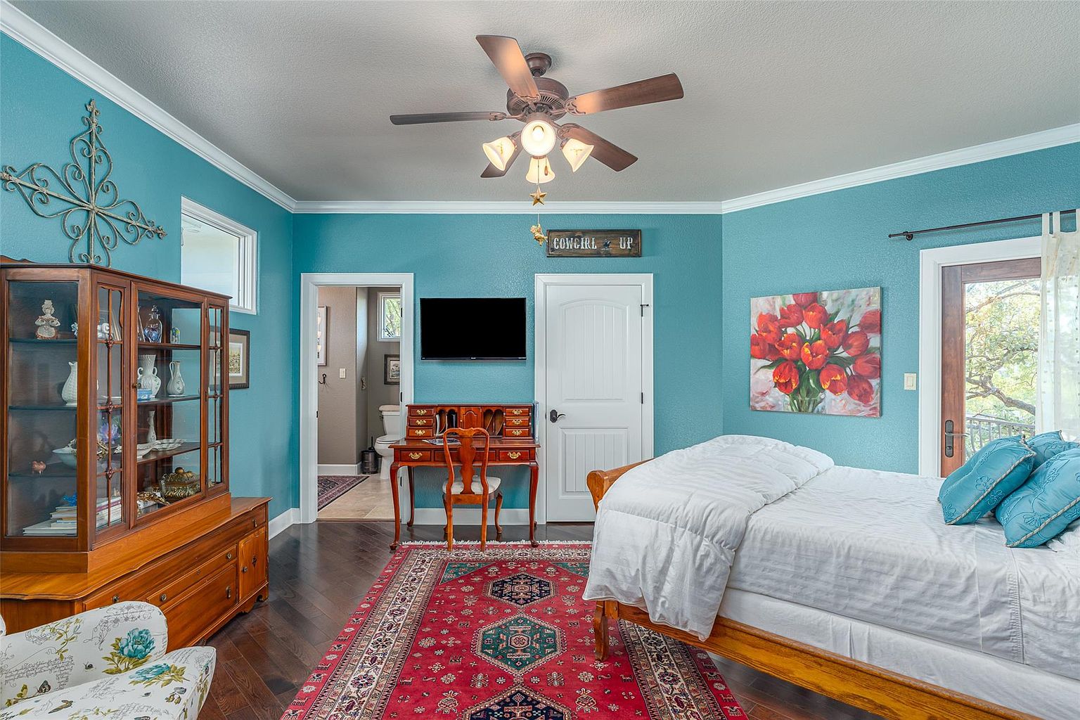 This is a cozy primary bedroom featuring a wooden bed frame with white bedding and blue pillows, a red oriental rug, and a wooden desk with a chair. The walls are painted a calming blue, complemented by white trim and a ceiling fan. A painting of red tulips adds a pop of color, and a doorway leads to an en-suite bathroom.