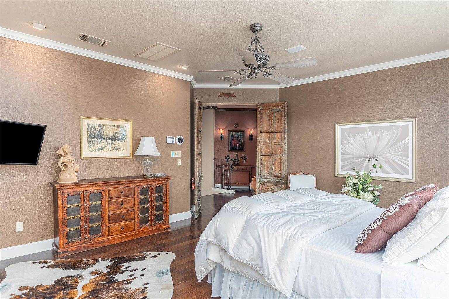 This is a primary bedroom featuring a large bed with white linens and decorative pillows. The room has a rustic-chic style with a wooden dresser, artwork, and a unique ceiling fan. An open doorway leads to another area, adding depth and interest to the space.