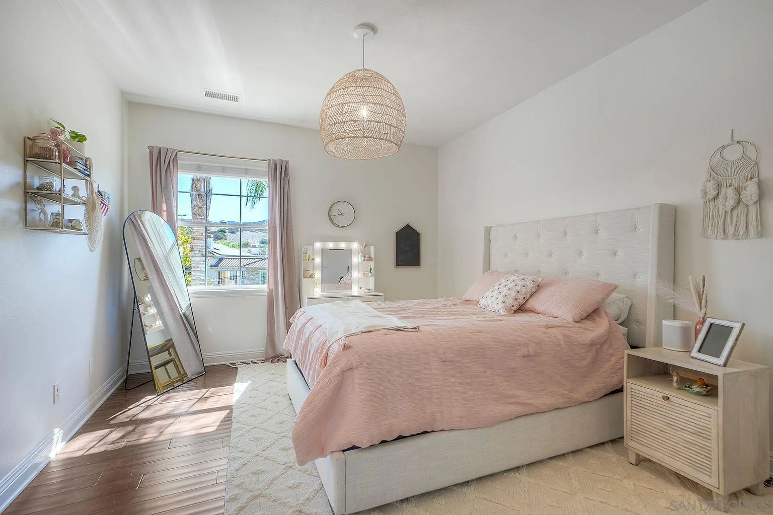 This is a well-lit bedroom featuring a bed with a light pink comforter and a tufted headboard. The room is decorated with a woven pendant light, a full-length mirror, and a macrame wall hanging, creating a cozy and stylish atmosphere. A vanity with a lighted mirror and a nightstand with decorative items add to the room's charm.