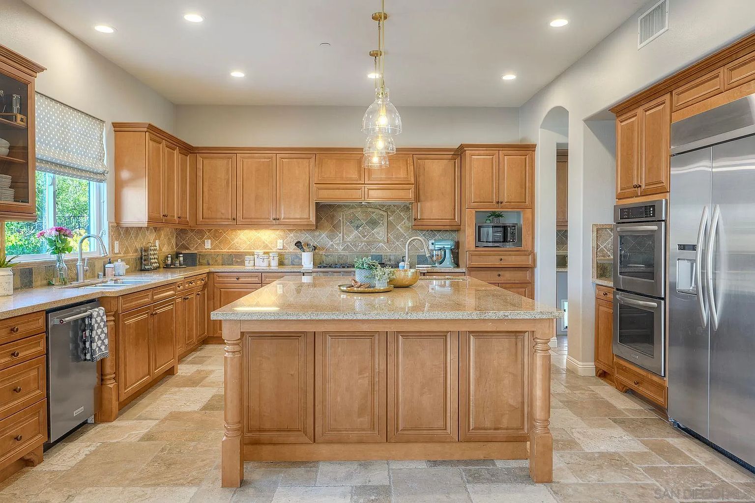 This is a well-lit kitchen featuring wooden cabinetry, stainless steel appliances, and a large island with a granite countertop. The kitchen has a traditional style with a decorative backsplash and pendant lighting above the island. The flooring is tiled, and the overall impression is one of warmth and functionality.