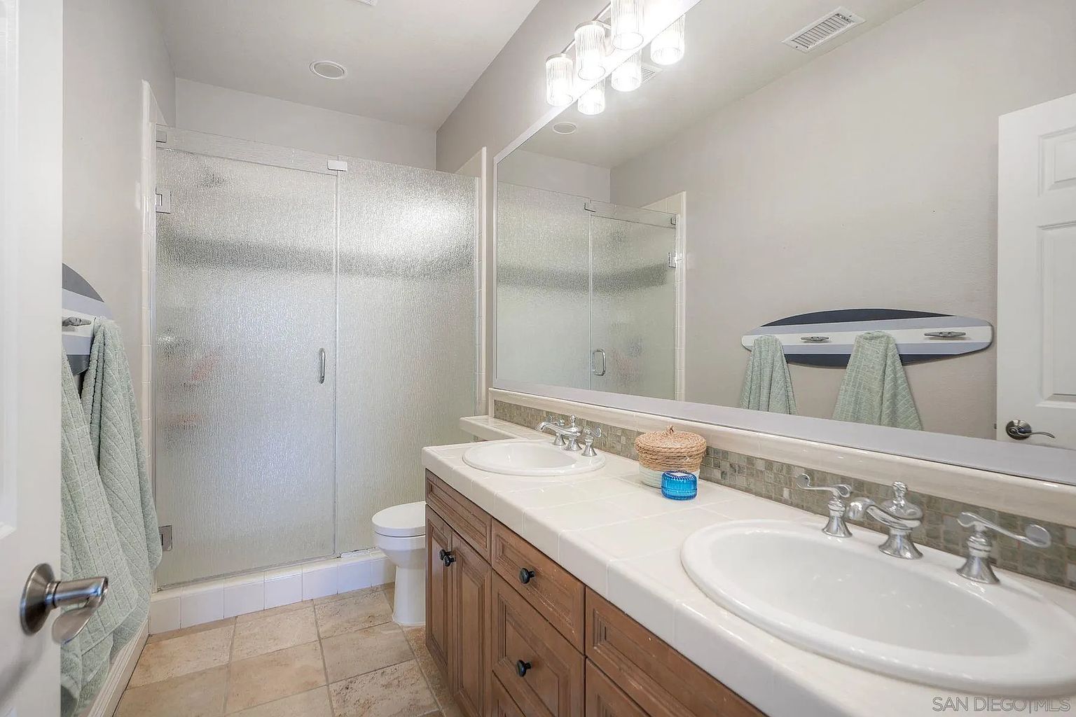This is a well-lit primary bathroom featuring a double vanity with white countertops and wooden cabinets. A large mirror reflects the vanity and a glass-enclosed shower. The flooring is tiled, and the overall impression is clean and functional.