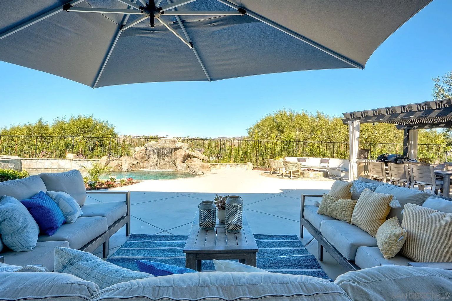 This image showcases a luxurious outdoor patio area, complete with comfortable seating arrangements, a stylish rug, and a view of a pool with a waterfall feature. A large umbrella provides shade, while a pergola with a dining area is visible in the background, creating an inviting and upscale outdoor living space perfect for entertaining or relaxation.