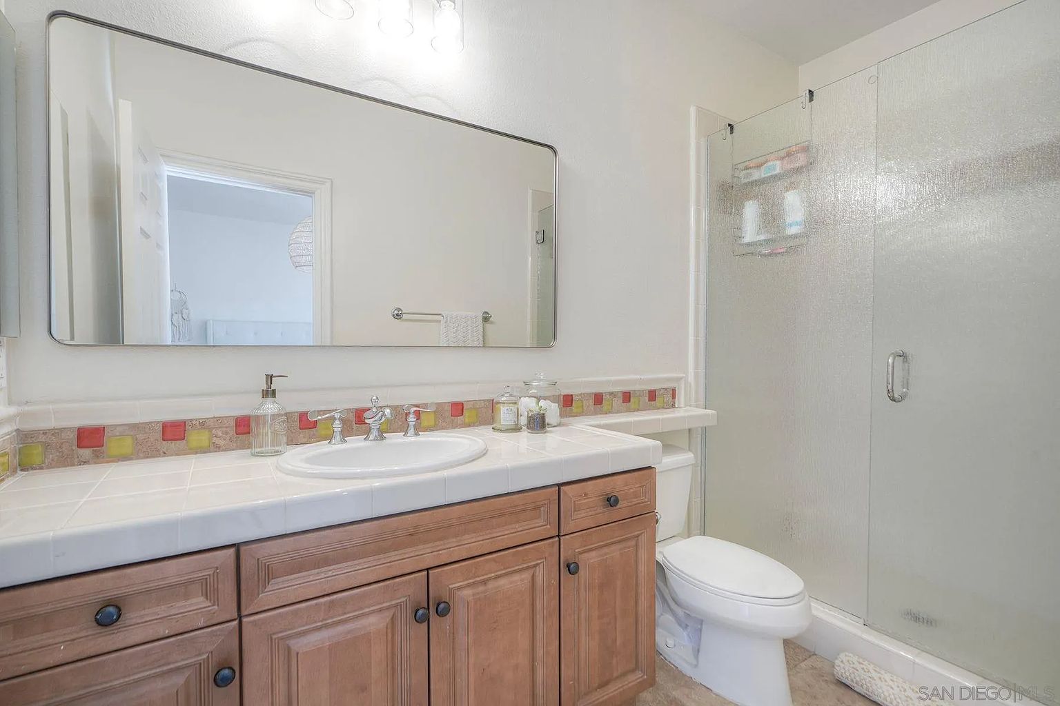 This is a bathroom featuring a vanity with a white countertop and wooden cabinets, a white sink, and a large mirror above the vanity. A shower with a frosted glass door is visible to the right, and a toilet is positioned next to the shower. The bathroom has a clean and functional design.