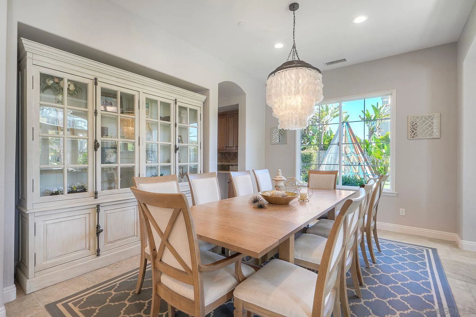 This is an inviting dining room featuring a large wooden dining table surrounded by upholstered chairs. A decorative chandelier hangs above the table, and a built-in hutch provides ample storage and display space. The room is well-lit with natural light from a nearby window, creating a warm and welcoming atmosphere.