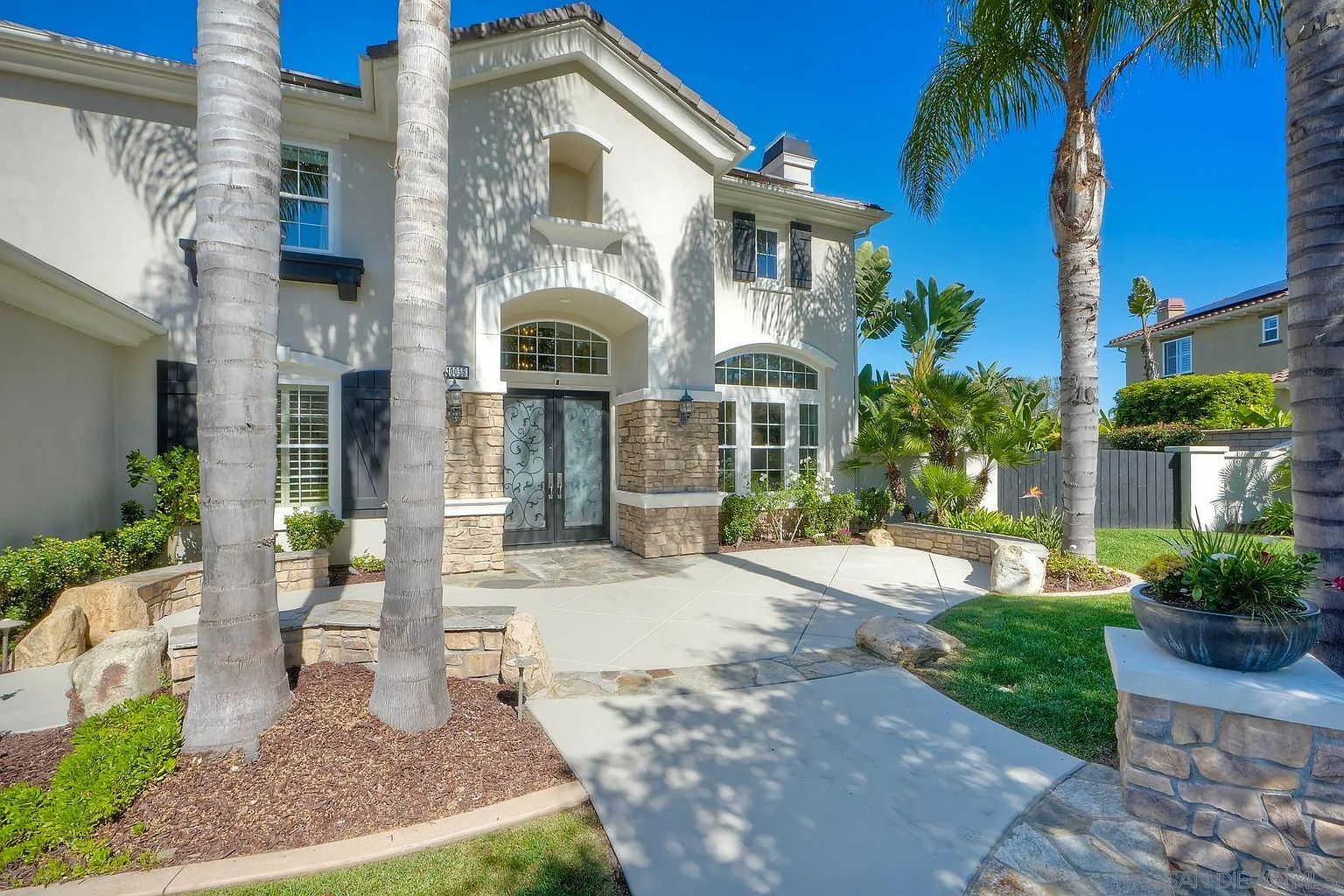 This is a front exterior view of a two-story house with a stucco facade and stone accents around the entryway. The house features a double-door entrance with decorative ironwork, arched windows, and dark shutters. Palm trees frame the house, adding to the property's curb appeal and creating a tropical feel.