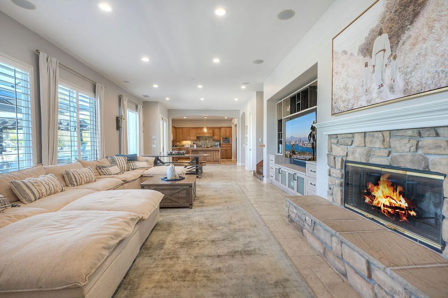 This is an interior shot of a living room that flows into the kitchen. The living room features a large sectional sofa, a stone fireplace with a mounted television, and a neutral-toned rug. The open floor plan creates a spacious and inviting atmosphere, perfect for entertaining or relaxing.