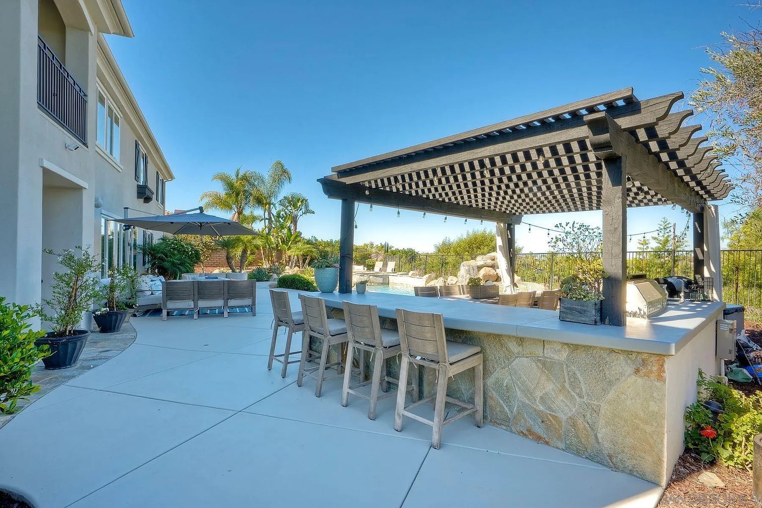 This image showcases a luxurious outdoor patio area, featuring a stylish pergola with a checkered pattern, a built-in bar with stone accents and bar stools, and comfortable lounge seating. The patio overlooks a pool area and lush landscaping, creating a relaxing and inviting atmosphere. The clear blue sky enhances the overall appeal of this outdoor living space.